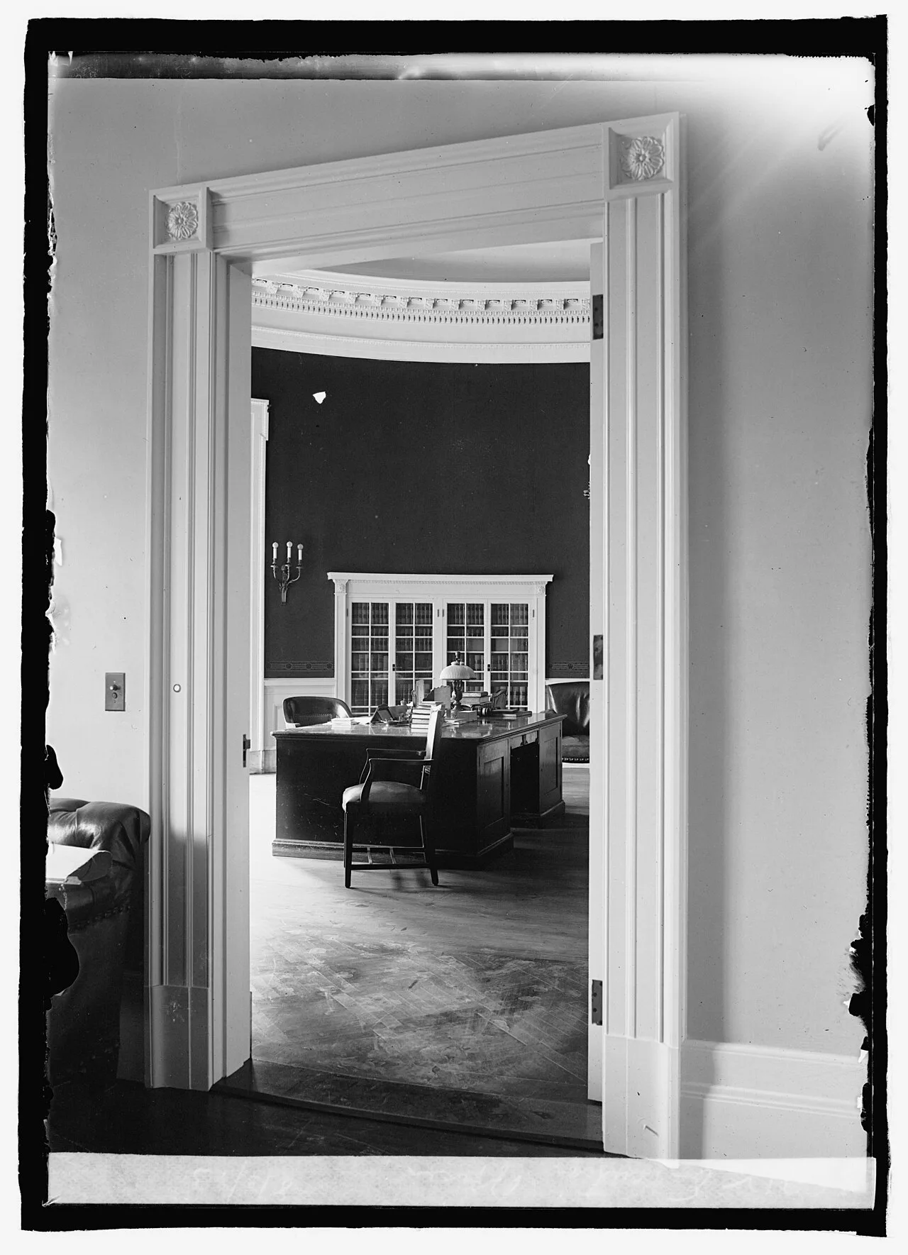 Foto en blanco y negro de un despacho oval visto desde una puerta, con escritorio, estanterías y suelo de madera