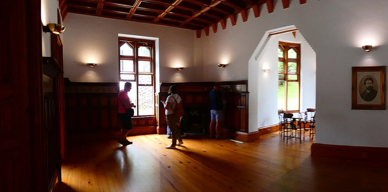 Cenador de El Capricho con zócalo alto de madera oscura, chimenea, ventanas ojivales y suelo de tablas largas de tono miel
