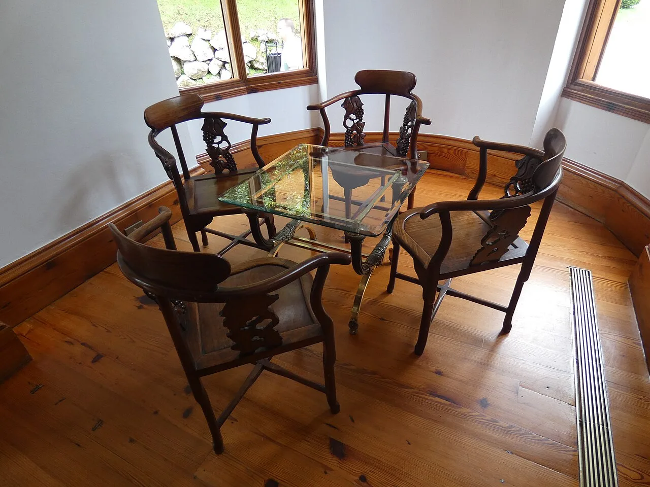 Galería del fumadero con mesa de cristal, sillas de madera tallada y suelo de tablas con nudos y una rejilla metálica junto a la pared