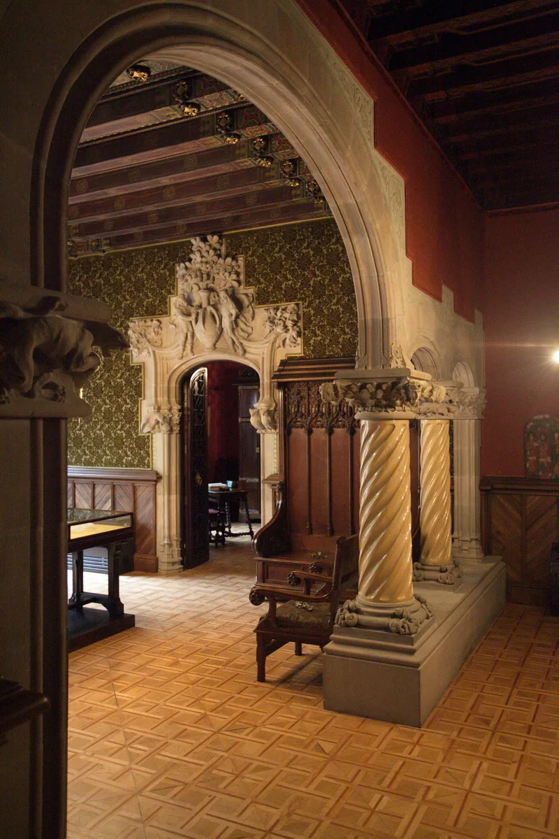 Parqué de losas cuadradas de tono miel que pasa bajo un arco de piedra junto a columnas salomónicas, hacia una sala de papel verde