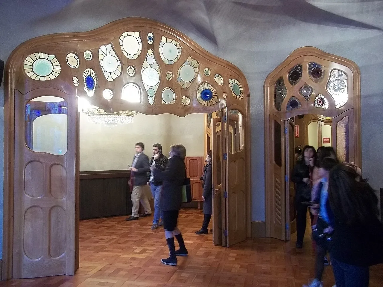 Puertas de madera con vidrieras de colores en el salón principal de la Casa Batlló y visitantes sobre el suelo de losetas de tablillas