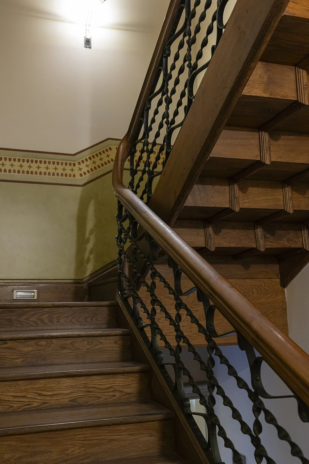 Tramo de escalera con peldaños de madera barnizada de canto redondeado, la estructura de madera del tramo superior vista por debajo y barandilla de barrotes de forja retorcidos