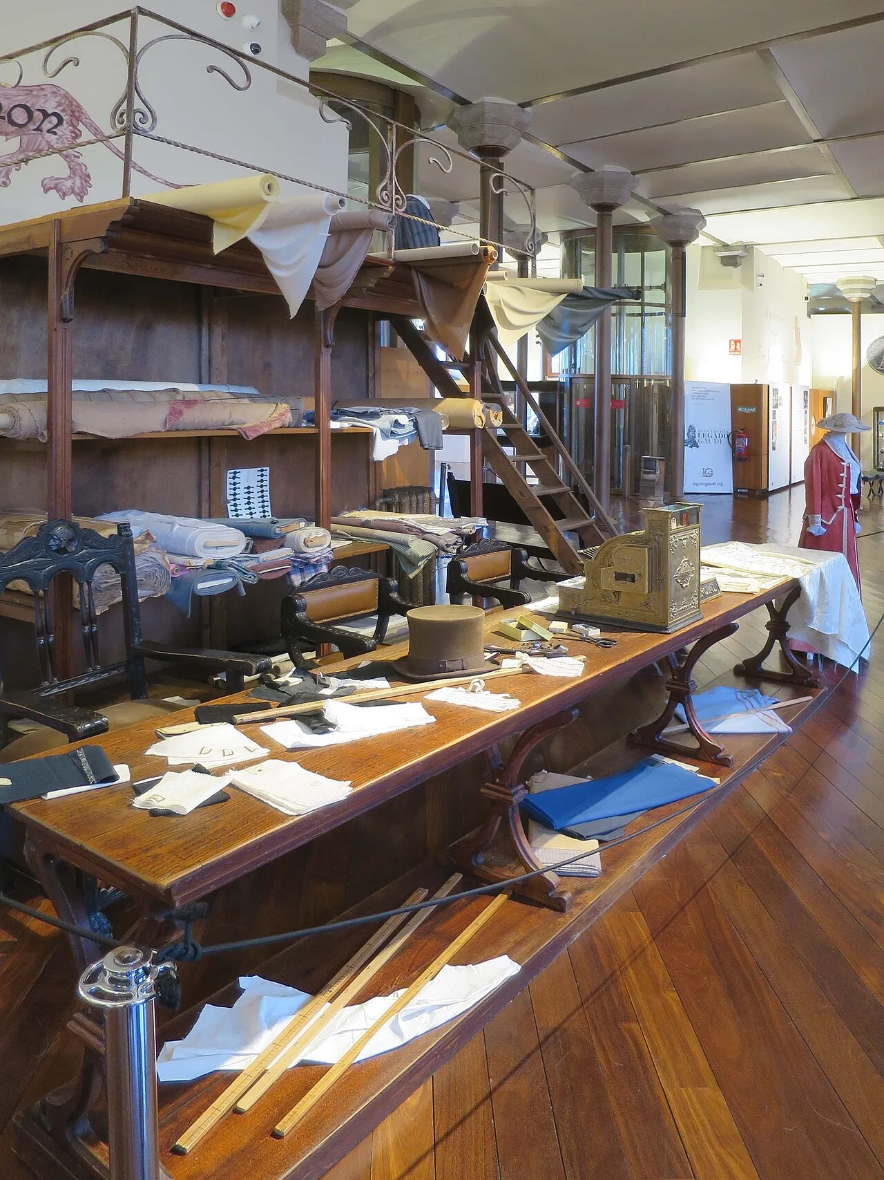 Recreación de una tienda de tejidos con altillo de madera, escalera de tablones, mostrador largo con telas, sombrero de copa y caja registradora sobre una tarima de tablas estrechas