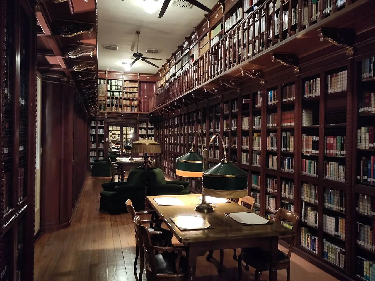 Biblioteca del Parlament balear con estanterías de madera de dos alturas, mesa de lectura con lámparas verdes y entarimado de tablas largas en tono miel